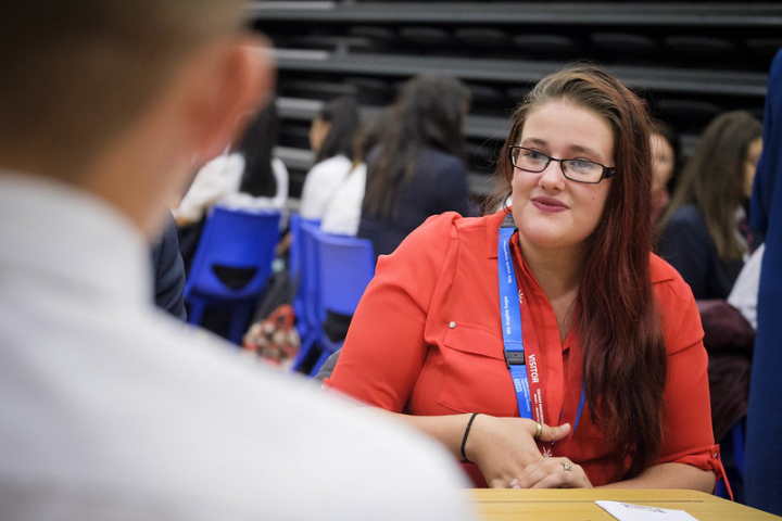 A female volunteer wearing a red shirt and blue NHS lanyard sits at a table with a student. Students show in the background sitting on blue chairs.